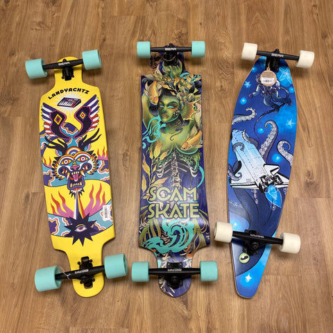 3 Longboards on the floor. The brand Scam Skate is in the middle and also on the right